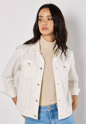 Chaqueta de mezclilla blanca con cierre de botón y dos bolsillos en el pecho, llevada sobre un suéter de cuello alto beige. Presenta mangas remangadas y jeans de color azul claro.