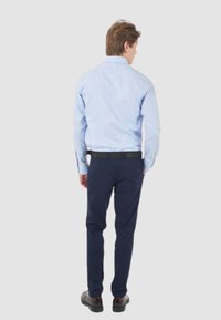 Camicia a maniche lunghe azzurro chiaro con taglio dritto, abbinata a pantaloni blu scuro e una cintura nera con un design a motivo. Le scarpe sono scure.