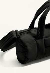 Svart tygväska med rektangulär form, utrustad med dubbla handtag, quiltad textur och en slät yta, accentuerad med en dragkedja med varumärkesdetalj.