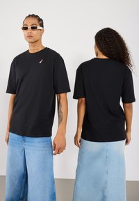 Svart bomulls t-skjorte med et lite rødt og hvitt brodert design på brystet. Korte ermer, rund hals, avslappet passform.