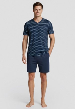Man staat blootsvoets, draagt een marineblauw kort mouwen V-hals overhemd met patroon en effen marineblauwe shorts tegen een effen achtergrond.