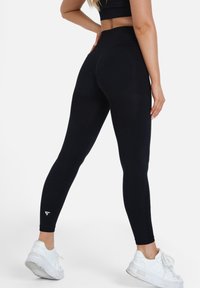 Schwarze, hochtaillierte Leggings mit enganliegendem Schnitt, strukturiertem Stoff und einem kleinen weißen Logodetail am Unterschenkel. Getragen mit weißen Sneakers.