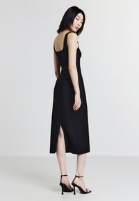 Vestito nero senza maniche con una silhouette aderente, di lunghezza media, spacco laterale e un design con schiena bassa. Indossato con sandali con tacco alto e fasce.