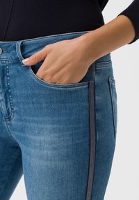 Modré džíny z denimové látky s úzkým střihem, s boční kapsou, kontrastním prošíváním a mírným zesvětlením. Ruka je opřená v kapse.