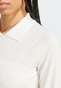 White long-sleeve polo shirt with a ribbed collar, featuring subtle embossed logo on the chest and smooth fabric texture.