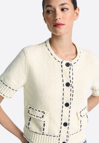 Kortärmelig crème cardigan met marineblauw stiksel, knopen aan de voorkant en twee opgestikte zakken. Getextureerde stof voegt een tactiele dimensie toe.