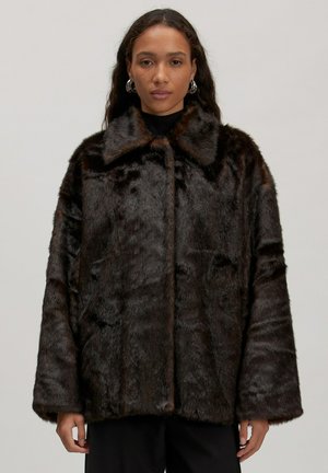 Manteau en fausse fourrure marron avec un large col, coupe ample et épaules tombantes. La surface présente une finition douce et texturée avec des variations de couleurs riches.