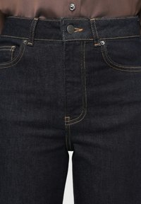 Vaqueros de mezclilla oscura con cintura alta, cuentan con costuras doradas contrastantes, cierre de botones y bolsillos delanteros. Textura suave y diseño clásico.