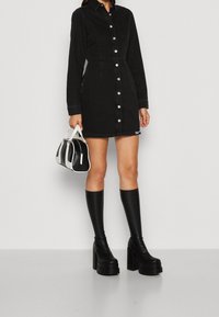 Černé džínové šaty košile s knoflíky vpředu, límečkem a přiléhavým pasem; nošeny s černými platformovými botami do půli lýtek a malou černobílou kabelkou.