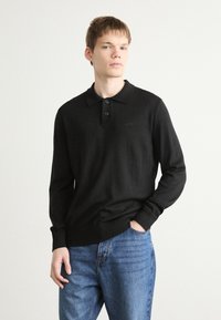 Schwarzes Langarm-Polohemd mit geripptem Kragen und Bündchen, ausgestattet mit drei Knöpfen und subtilen Logodetails auf der Brust. Kombiniert mit blauen Jeans.
