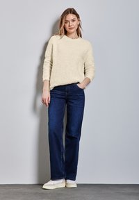 Kremowy, puszysty sweter dzianinowy z długimi rękawami, zestawiony z ciemnoniebieskimi dżinsami o szerokich nogawkach i jasnymi, strukturalnymi trampkami.
