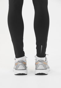 Leggings deportivos negros con acentos reflectantes y un diseño ajustado, combinados con zapatillas de running grises que presentan detalles en naranja y negro.