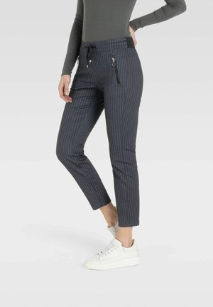 Femme portant un pantalon court gris à fines rayures avec des poches zippées et des baskets blanches, debout sur un fond clair uni.