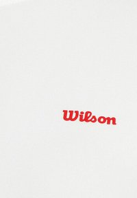 Tela blanca con una superficie texturizada que presenta un logo rojo de "Wilson" en el centro. El logo es audaz, curvilíneo y se muestra de manera prominente.