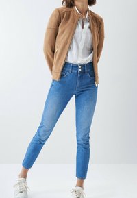 Hnědá semišová bunda s detaily na ramenou, bílá košile na knoflíky a světle modré úzké džíny. Outfit doplňují tenisky.