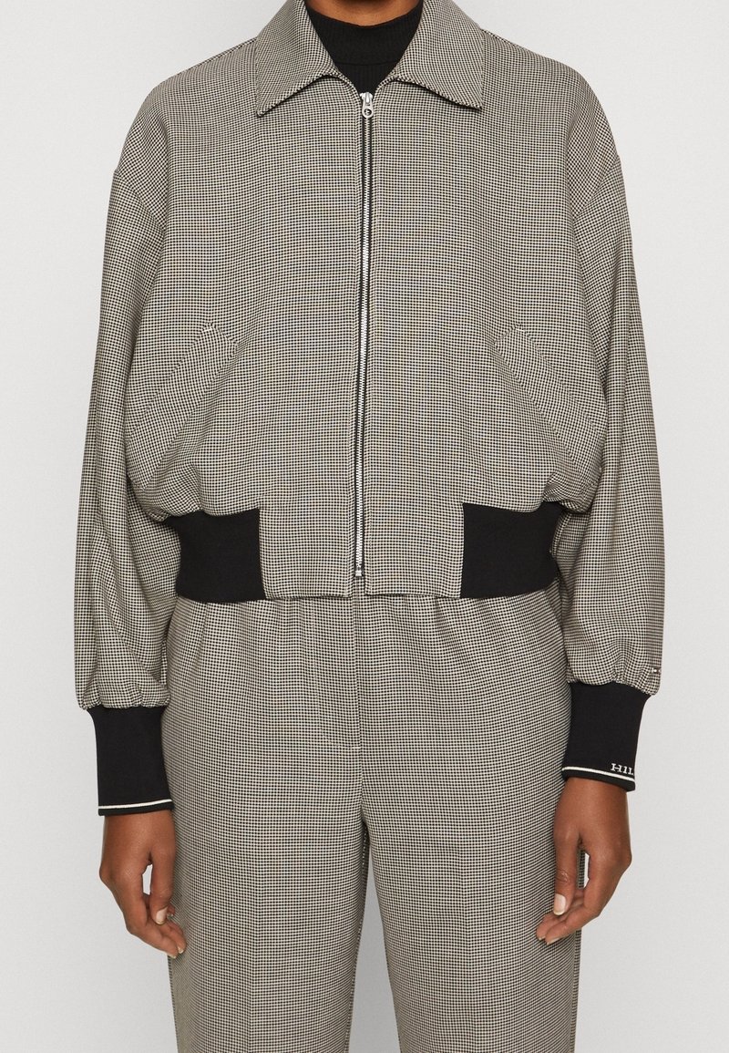 Personne portant une veste zippée à petits carreaux gris et noirs avec des poignets et une ceinture noirs élastiques, associée à un pantalon à carreaux assorti.