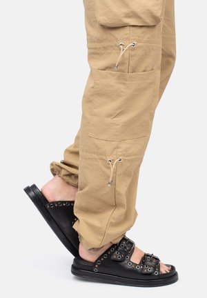 Pantaloni cargo beige con polsini regolabili e molteplici tasche, indossati con sandali neri con accenti borchiati e cinturini regolabili.