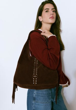 Jeune femme en pull bordeaux et jean bleu portant un grand sac à franges en daim marron avec des clous en argent sur l'épaule.