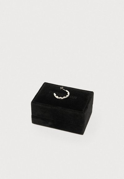 Ασημένιο μισοφέγγαρο σκουλαρίκι με σχεδιασμό με κυματιστό περίγραμμα που ακουμπά σε κλειστό ορθογώνιο κουτί κοσμημάτων από μαύρο βελούδο.
