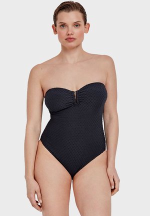 Traje de baño negro sin tirantes de una pieza que presenta un tejido texturizado, un frente fruncido y un detalle de ojal en el centro. Aberturas para las piernas de corte alto.