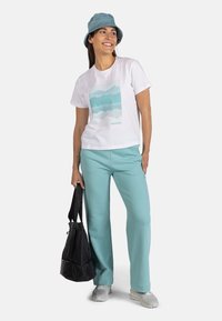 Camiseta blanca con un gráfico de montaña azul, combinada con pantalones verde claro y un bolso negro. El modelo lleva un sombrero de cubo azul y zapatillas grises.