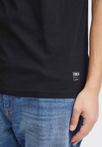 T-shirt en coton noir avec un col rond, des manches courtes et une petite étiquette de marque à l'ourlet. Porté avec un jean en denim bleu.
