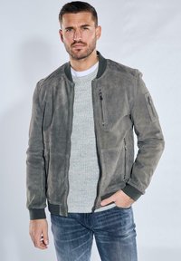 Graue Lederbomberjacke mit gerippten Bündchen und Kragen, mit Reißverschlusstaschen, getragen über einem hellgrauen gerippten Pullover und blauen Jeans.