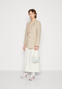 Μπεζ σακάκι με χρυσά κουμπιά, μπεζ τοπ, λευκή τζην φούστα, τσάντα με σχέδια μπλε και λευκό, αθλητικά sneakers με μεταλλικές λεπτομέρειες.