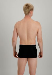 Short de bain noir avec un logo sur la taille, design ajusté et tissu lisse, porté par une personne de dos face à l'appareil photo.