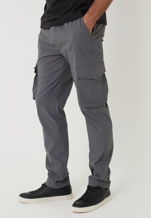 Mann trägt graue Cargohosen und schwarze Sneakers, steht mit einer Hand in der Tasche vor einfarbigem Hintergrund.