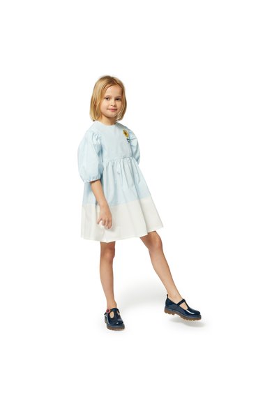 Robe en coton bleu clair avec des manches bouffantes, une taille froncée et un ourlet blanc. Présente un accent de broderie florale. Portée avec des chaussures navy.