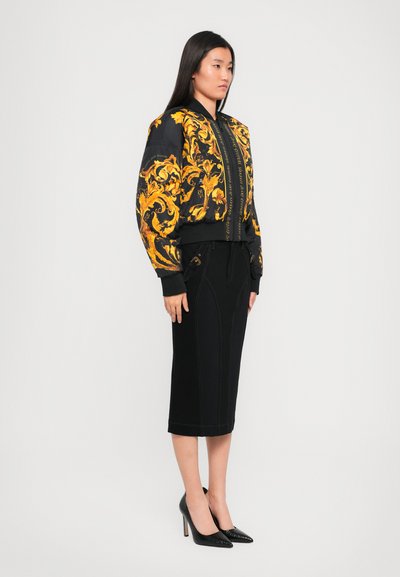 Veste bomber noire avec des motifs jaunes ornés, associée à une jupe noire longueur genou et des escarpins noirs à talons pointus. Texture lisse, design ajusté.