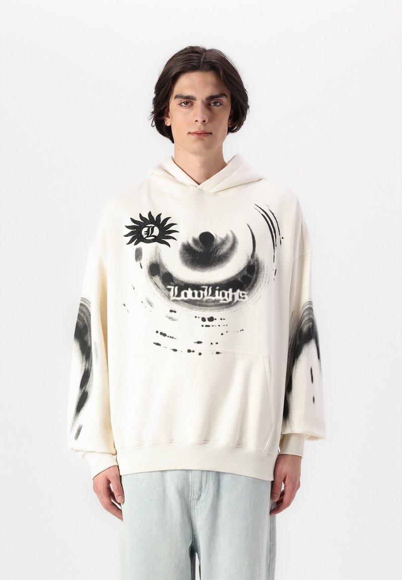 Hettegenser i krem med svarte grafiske mønstre, inkludert et øye og tekst. Myk stoff, avslappet passform og kengurulomme. Lys blå jeans.