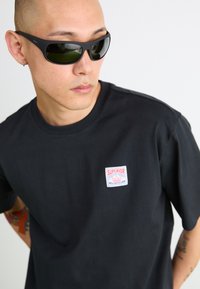 T-shirt noir en coton avec une petite étiquette "SUPERIOR" rouge et blanche sur la poitrine. Lunettes de soleil noires avec des verres verts.