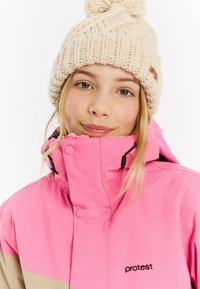 Rosa wasserdichte Jacke mit hellbraunen Akzenten, hohem Kragen und einem Reißverschluss vorne. Kombiniert mit einer cremefarbenen gestrickten Mütze mit einem Pompon oben.