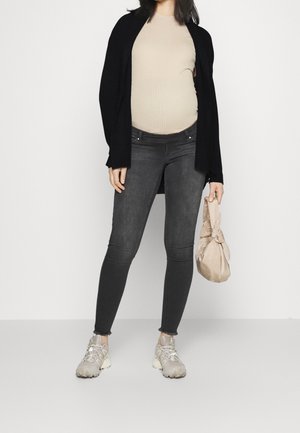 Mujer embarazada con top beige, cárdigan negro, jeans ajustados oscuros, zapatillas grises, sosteniendo una bolsa de tela atada de color beige claro.