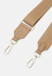 Béžový nastavitelný popruh na tašku s embosovaným textem, koženými konci a stříbrnými kovovými otočnými karabinami na bílém pozadí.