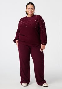 Zelfverzekerde vrouw in een bordeauxrode sweater met parelversieringen en bijpassende broek, gekleed in witte schoenen met studs, staande tegen een witte achtergrond.