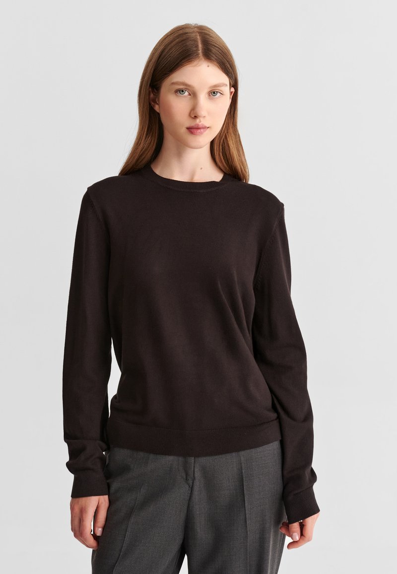 Jeune femme aux cheveux bruns et raides portant un pull noir à manches longues et un pantalon gris sur un fond clair uni.