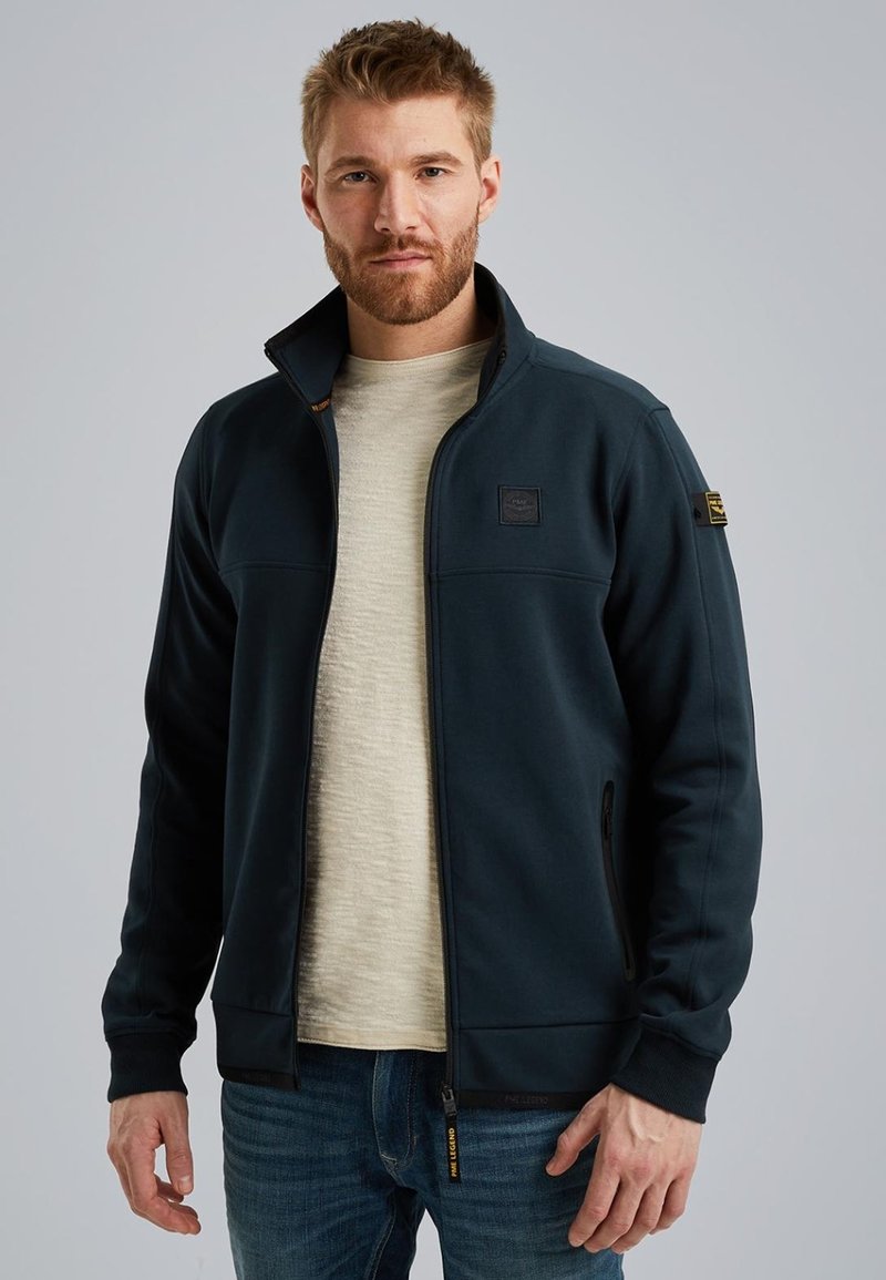 Donkerblauwe zip-up jas met hoge kraag, gemaakt van zachte stof; heeft twee zijzakken en een klein embleem op de borst en mouw.