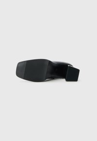 Chaussure à talon noir avec un talon en bloc rectangulaire, présentant une surface lisse et une semelle en caoutchouc noir texturé avec des rainures horizontales.