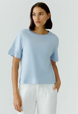 Vrouw met kort donker haar, gekleed in een lichtblauw shirt met korte mouwen en witte broek, staand met één hand in de zak, kijkend naar links.