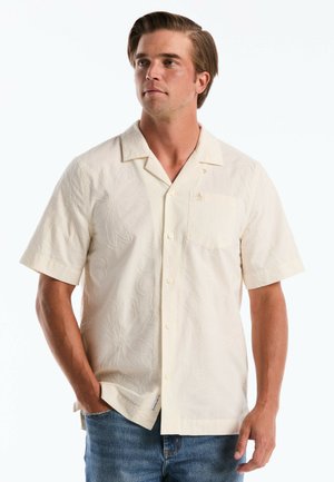 Man in een crèmekleurig overhemd met korte mouwen, met bloemenborduursel en blauwe spijkerbroek, handen deels in de zakken, kijkend naar links.