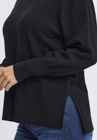 Fekete kötött pulóver ribbed aljjal és mandzsettákkal, oldalsó hasítékokkal. A textúrája puha, a fazon laza. Kék farmernadrággal viselve.