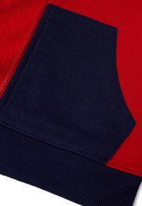 Červená textilie s tmavě modrým bavlněným kapsou, která má vroubkovaný lem a šicí detaily. Barvy výrazně kontrastují a zdůrazňují jednotlivé části.