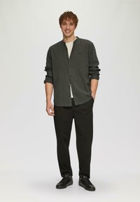 Camicia button-up verde scuro con colletto alla cinese, abbinata a pantaloni neri e sneakers nere, in piedi su uno sfondo neutro.