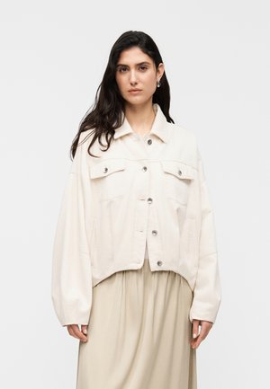 Gekürzte cremefarbene Jacke aus weichem Stoff, mit zwei Brusttaschen, silberfarbenen Druckknöpfen und lässigen Ärmeln, getragen über einem hellbeigen Rock.