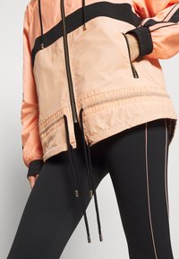Chaqueta bomber de color melocotón con acentos negros, bolsillos con cremallera, puños elásticos y cordones. Leggings negros con franjas laterales rosas.