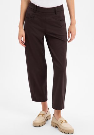 Femme portant un pantalon court marron foncé avec poches avant et des mocassins beiges avec des ornements dorés sur fond blanc.