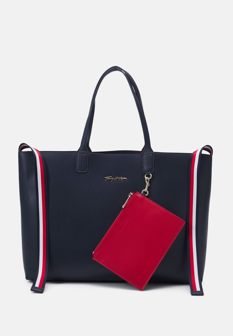 Sac fourre-tout marine avec double poignée, bandoulière rayée rouge-blanc-bleu, logo doré et pochette zippée rouge attachée.